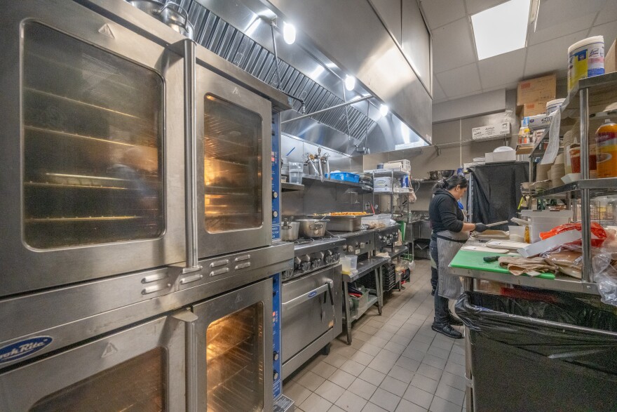 A woman wearing a black long sleeve shirt and a grey apron stands in a commercial kitchen. She is holding a knife to cut an ingredient for a prepared food product. In the kitchen, stacked ovens are visible as well as countertops with supplies.