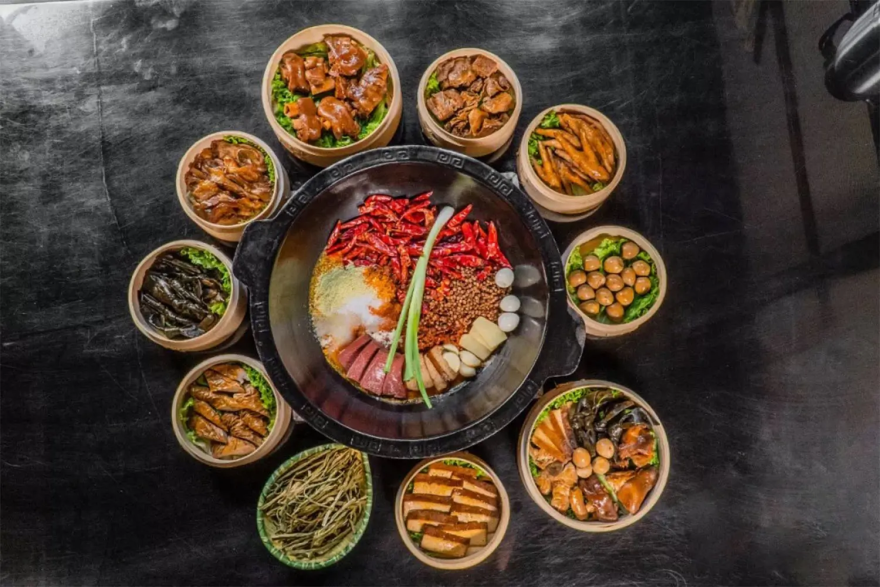 A large grey-colored stone dish containing a variety of meats and vegetables sits in the center of an assortment of round, light brown-colored steamer baskets, each containing different types of food on a black surface.