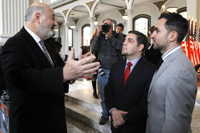 Three men in talking to each other. Rob Reiner is on the left with hands out in expression as he speaks with two younger men in suits on his right. Behind them is a photographer with a camera pointing towards them and to the far left are a row of American flags.