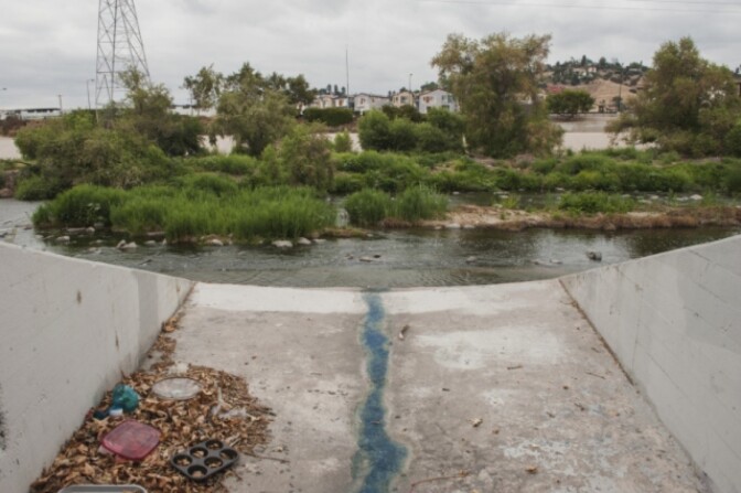 A storm drain along the LA River in the Frogtown neighborhood is cluttered with household trash and items, including what appears to be a muffin tin and a red lid. 