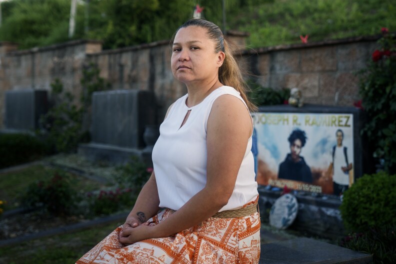 A woman wearing a white top and orange printed skirt sits in front of a tombstone with photos of a young man and the name Joseph R. Ramirez printed on it