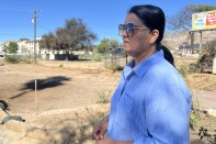 A woman with medium dark skin tone and blue shirt looks to the distance in front of an empty dirt lot.  