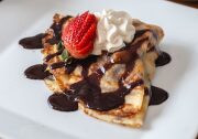 An image of a crepe sits on a white plate. The crepe is a light brown color and is covered with a dark brown chocolate sauce, a piped dollop of whipped cream and half a strawberry