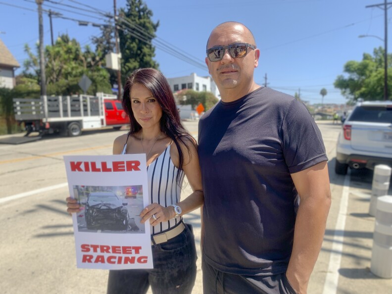 A woman in a white spaghetti strap tank top with thin black stripes holds a sign that says "Killer Street Racing" in red letters with an image of a crushed car front. A man in a navy blue shirt stands to her left.