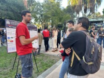 A man with light skin tone stands before a scientific research poster in a courtyard. In front of him, three other people listen as he speaks. In the background, people pause to look at other posters. 