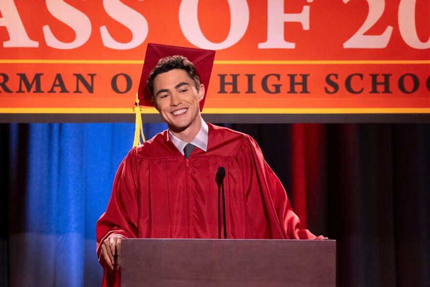 An actor playing a high school senior wears a red graduation cap and gown and stands in front of a podium smiling.