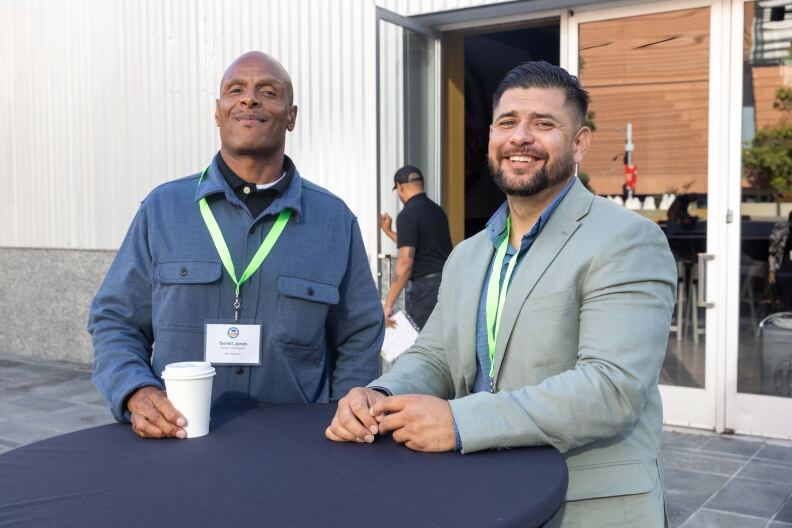 A bald dark skinned man wearing a blue shirt and an ID around his neck is standing next to a light skinned man with dark hair who is wearing a light greey jacket. They are both looking at the camera and smiling