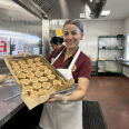A woman wearing a red polo, apron and hair net holds a baking sheet with freshly rolled cinnamon rolls.