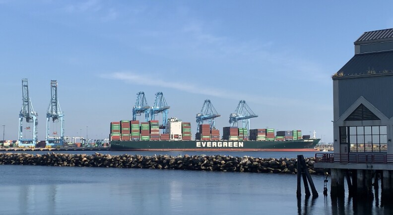 A cargo ship sits in the distance in the harbor waiting to unload containers of goods. It's lined with green and rust-colored steel boxes.