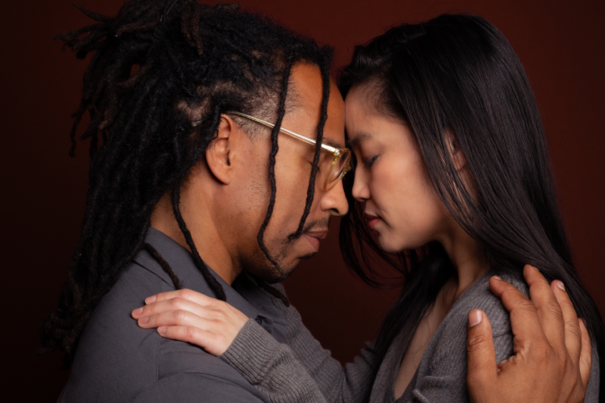 A man and woman embrace with their hands resting on each others' shoulders in front of a burgundy colored background. They are resting their foreheads against each other with their eyes closed. The main has dark skin tone with a thin black goatee and dreadlocks. He is wearing a gray collared shirt, with eyeglasses with clear plastic yellow frames and has his eyes closed. The woman is of Asian ethnicity with light skin tone and black hair. She is wearing a long gray sweater. She has shoulder length black hair and reddish lipstick. 