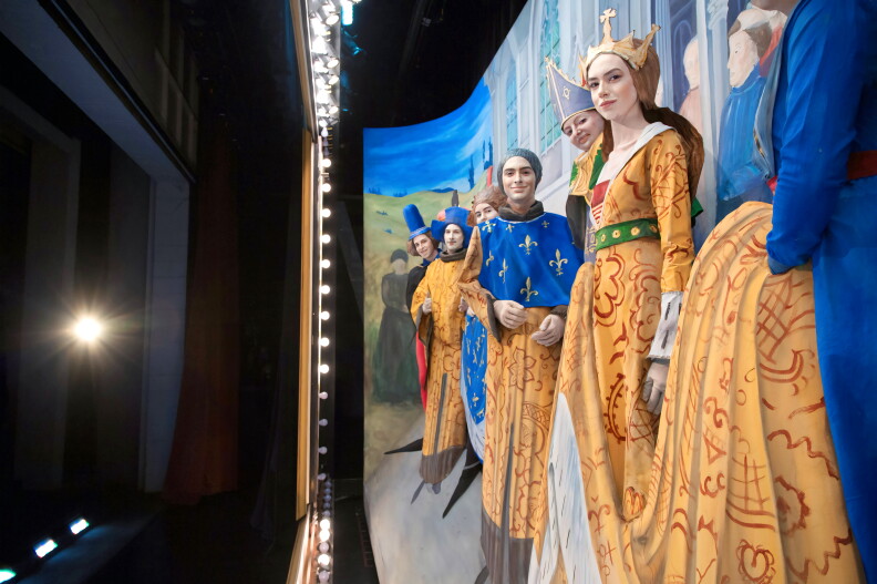 A line of women and men stand in front of a backdrop wearing medieval clothing and pointy black shoes.