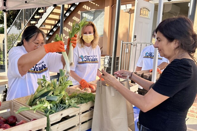A female presenting person puts vegetables into a paper bag held by a female presenting person.