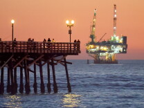 A pier is in the foreground. Behind is an offshore oil and gas platform. The sun is setting.