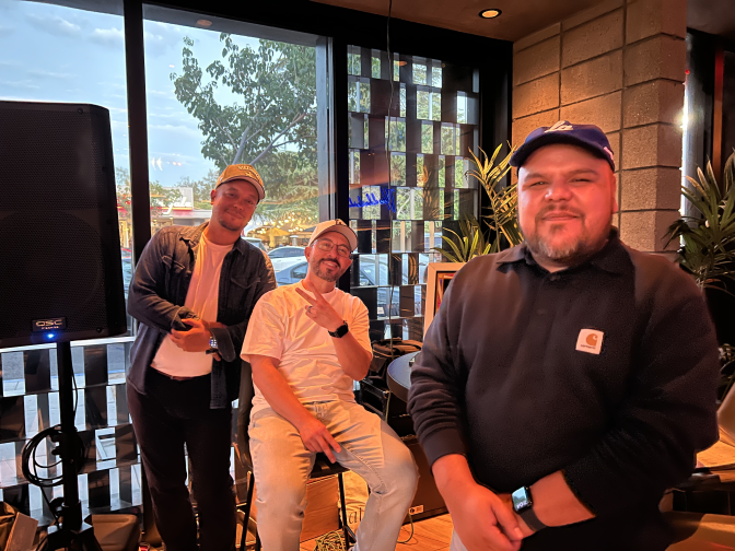 Three men with hats pose for a picture. The one on the far left wears a white shirt and a blue jacket. The one in the middle wears a white shirt and is throwing a peace sign. The one on the right edge is holding his hands crossed in front of his body.