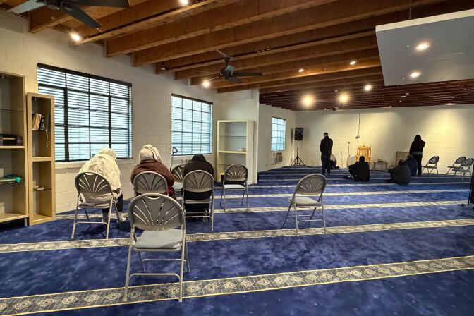 A well lit space with wooden beams in the ceiling a thick blue carpeting. Some people are engaged in the Muslim prayer. 