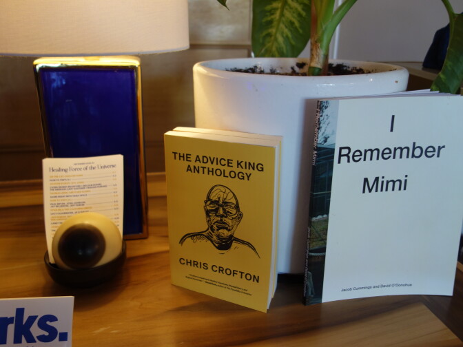 Two paperback books on a wooden shelf beside a white planter: “The Advice King Anthology” by Chris Crofton, with a yellow cover and a line-drawn portrait of a bearded man, and “I Remember Mimi,” with a white cover.