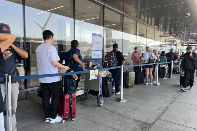 Passengers holding on to wheeled suitcases stand in a line behind blue tape. Some passengers carry backpacks. 