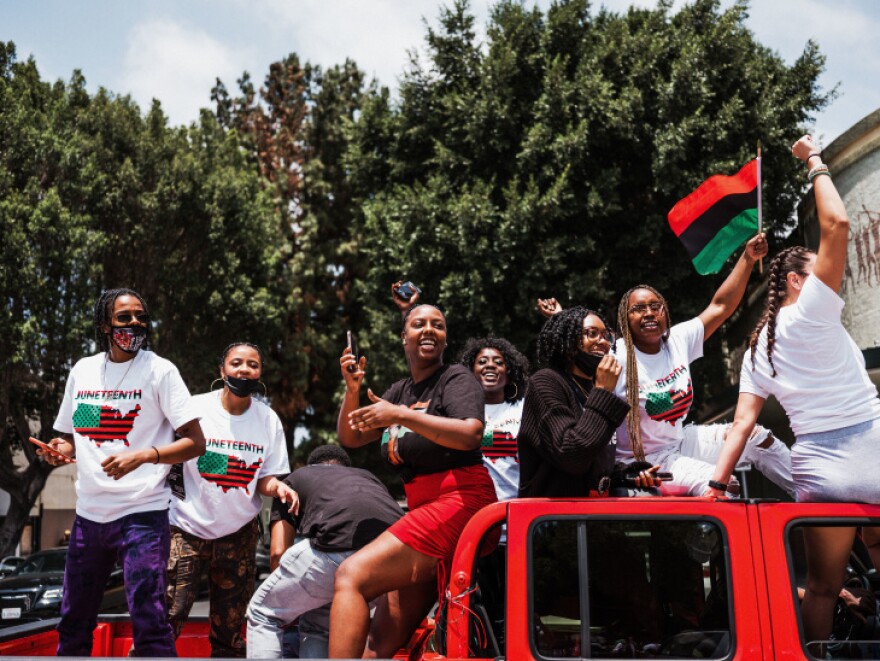 A group of Black men and women smile and wear shirts that read "Juneteenth" as they ride in a red truck and wave flags.