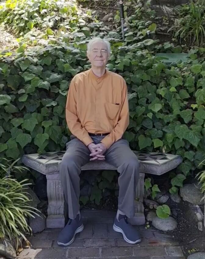 A man with gray hair and light skin in an orange shirt meditates while sitting on a bench, surrounded by green leaves.