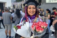 A woman with medium skin tone and long burgundy hair smiles while holding two bouquets of flowers. She is clad in a black cap and gown and drenched in colorful stoles and ribbons. 