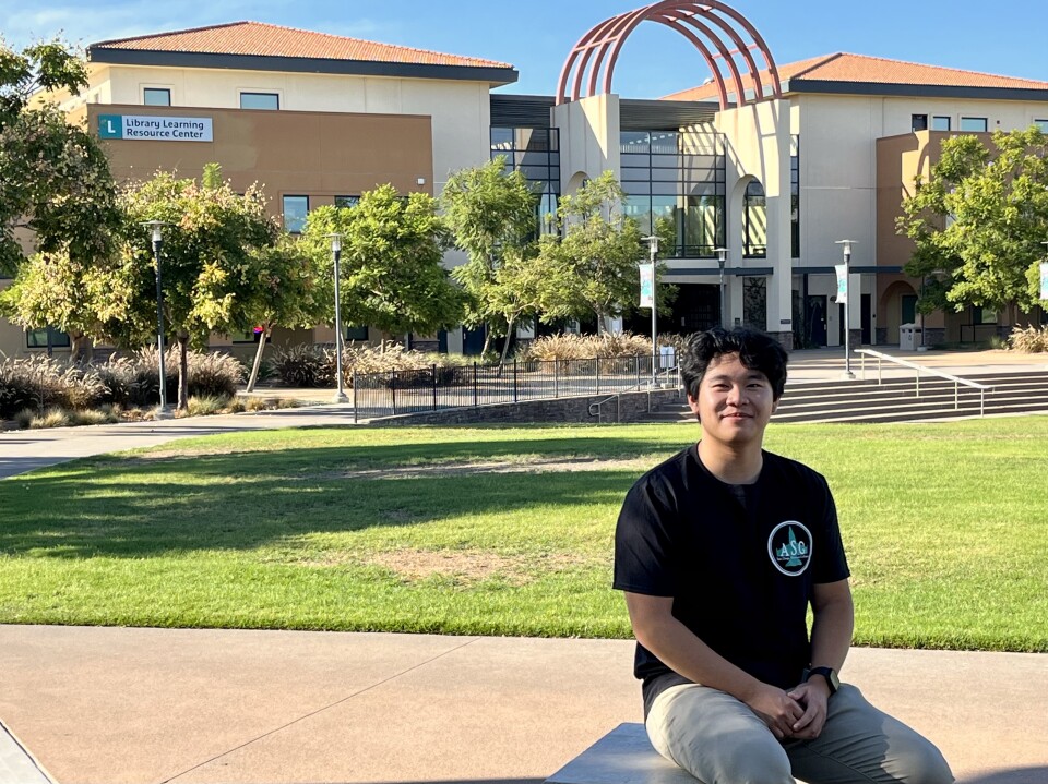 A young man in a black shirt sits on a bench in front of a lawn at his college campus. There is a large building in the background.