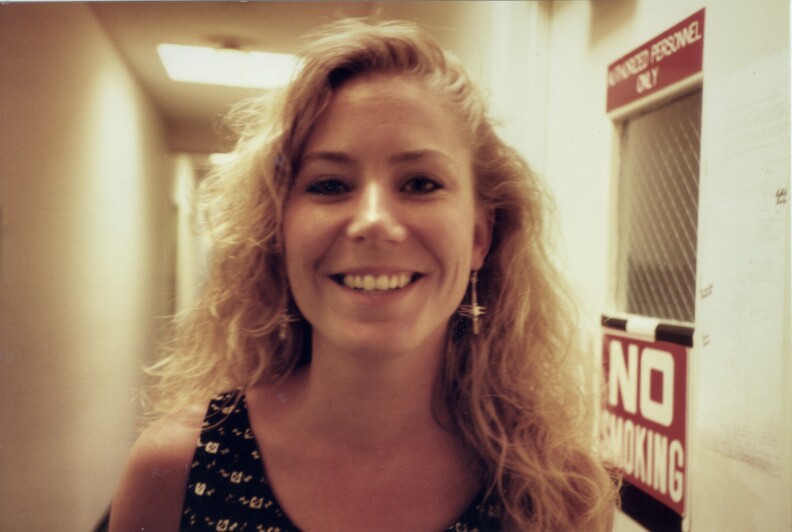 MARS FM DJ Holly Adams stands in a hallway smiling. She has blonde curly hair and is wearing long earrings. She's standing next to a door with two signs. One reads 'Authorized Personal Only' above a door window and the other reads "No smoking"