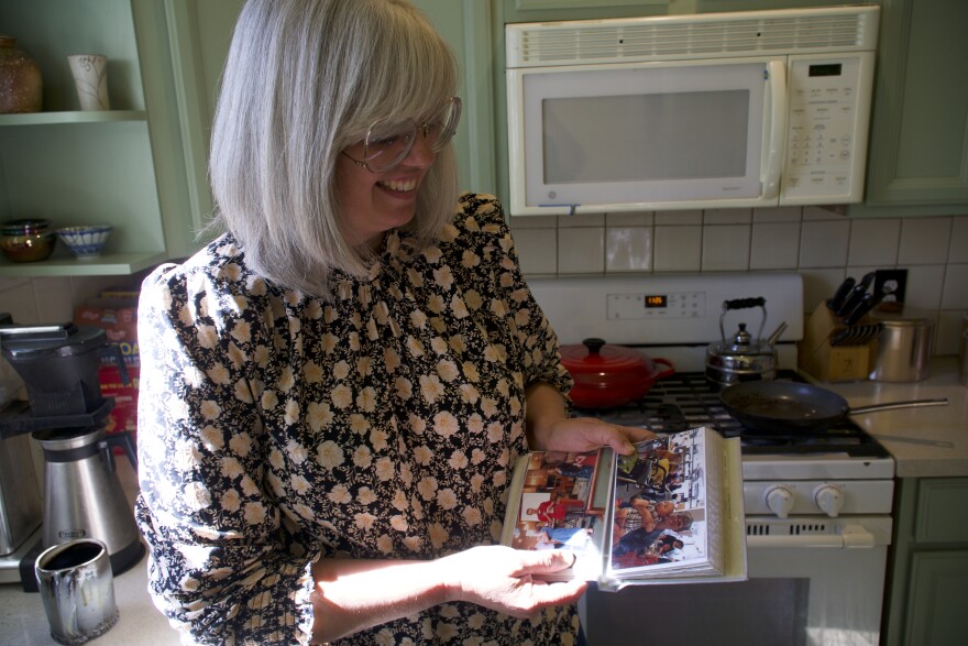 A light skinned woman standing in her kitchen smiles as she holds a photo album.