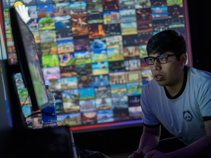 A player with medium-tone skin, wearing glasses and a T-shirt with a panda design, is focused holding a gaming control while looking at a screen. Behind the background is a projected image out of focus.