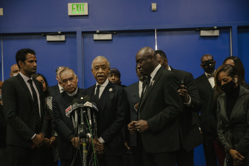 Four men, dressed in black, stand in front of a collection of microphones. Other men and women are in the background and around them.
