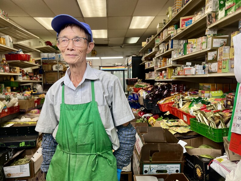 A faintly-smiling older Asian man wearing glasses, a blue cap, and a green apron stands inside a store, its shelves packed with boxed goods.