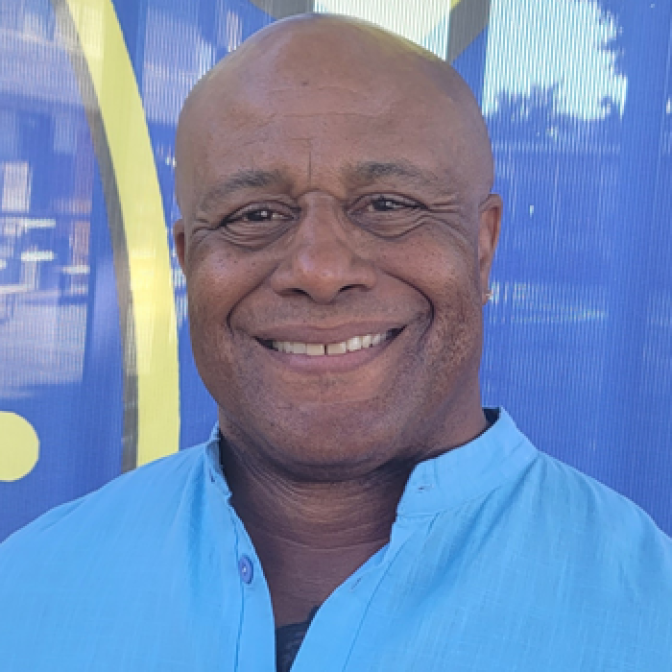 A man wearing a bright blue shirt is standing in front of a blue background, and smiling for the camera.  