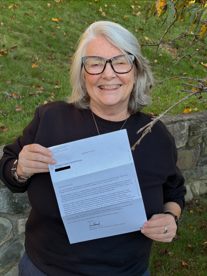 A woman with shoulder-length white-and-gray hair wears a black sweater and cat-eye glasses. She holds a letter with printing on it in her hands.