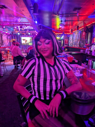 A drag performer wearing a soccer jersey sits at a bar, smiling.