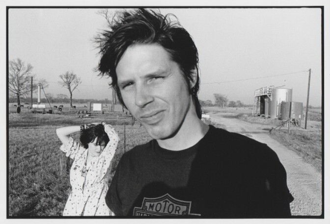 A man with short hair and a woman in a flowery dress behind him in a countryside setting.