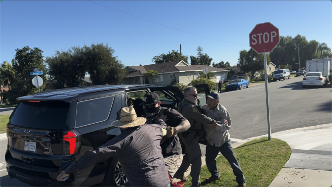 Light-skinned men with dark sunglasses—one with a Border Patrol uniform and one in plainclothes— grapple two gardeners with medium skin tone. Behind them, a black SUV is parked with its doors open. Across the suburban street, a bystander records the scene with their phone.