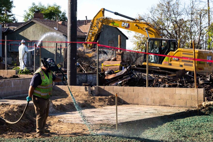 A person wearing a yellow safety vest and black helmet sprays a dark green liquid from a hose onto a piece of property. Behind the person is a tractor and a person in a white protective suit spraying water onto a property.