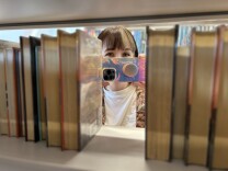 A light-skinned woman with medium-dark hair and bangs holds up a phone to use as a camera. She is peering from behind a shelf lined with books. 