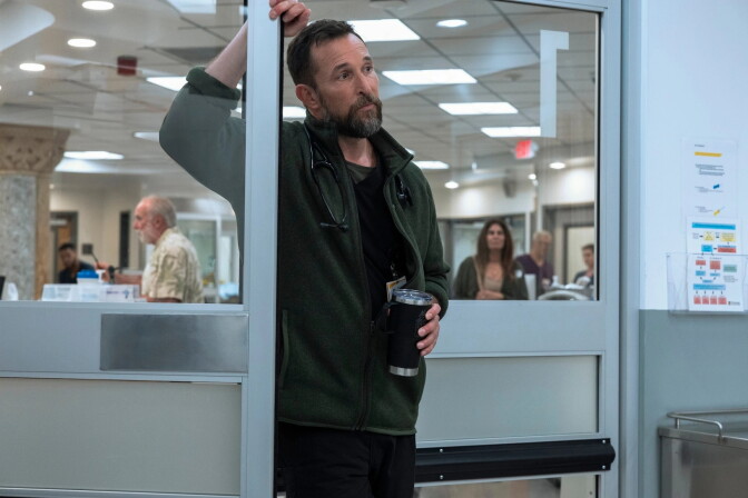 A man holding a black coffee cup leans on a the frame of a glass door, in a hospital. He is wearing a green long sleeve top and a stethoscope around his neck.