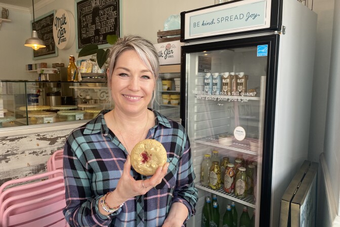 A woman with grey hair smiles while holding a large cookie inside a decorated bakery with signs reading "Milk Jar Cookies."