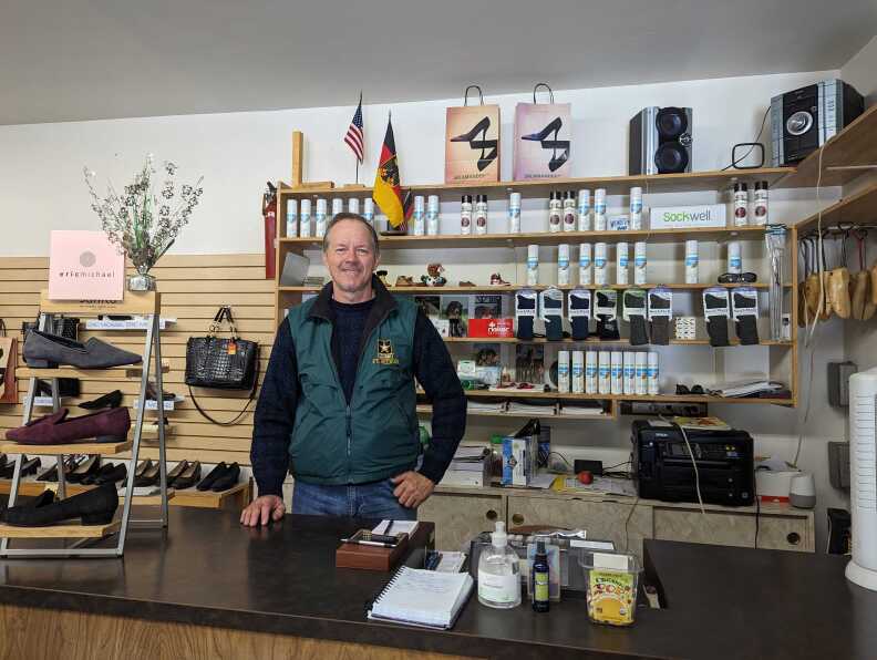 A light-skinned man stands behind a counter with surrounding. shoes and other products on shelves.