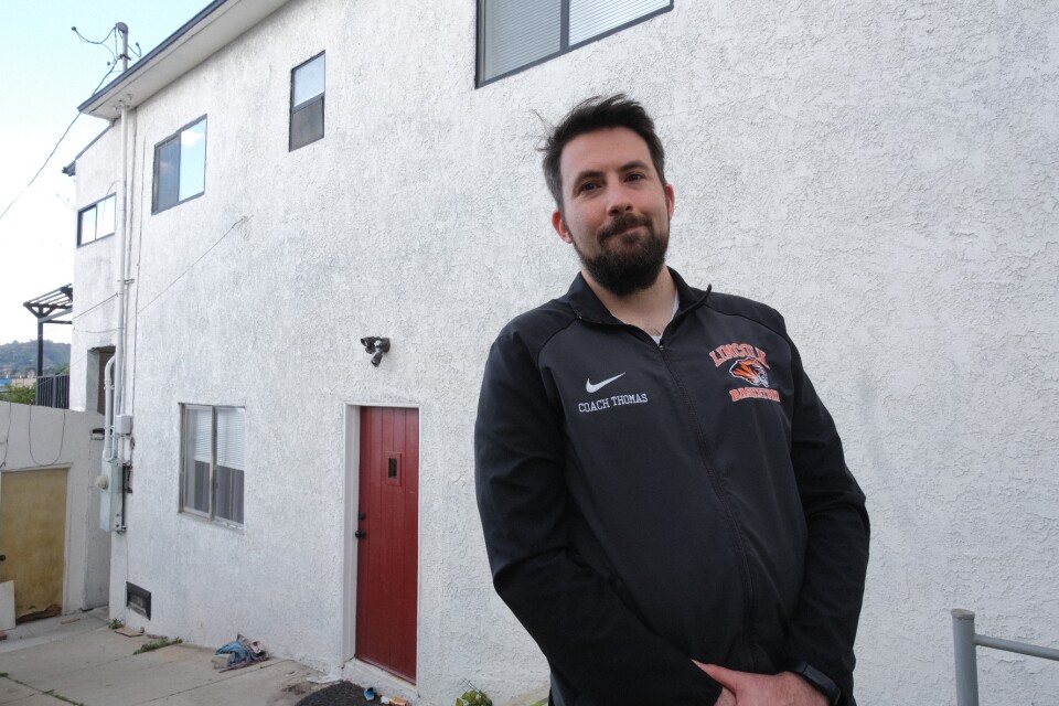 A man with light-tone skin and a beard and mustache stands outside a duplex with a red door. His jacket reads: Coach Thomas and has a Lincoln High emblem.