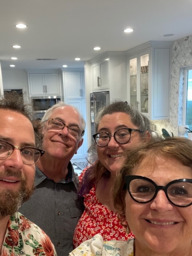 A group of light skin people pose for a photograph together. There are two men and two women. All of them are wearing glasses. The older man in back has white hair. The man closer towards the front has a beard and facial hair and is wearing a floral printed button up shirt. The woman in the back is wearing a red dress. The woman in the front has brown hair and is smiling. 