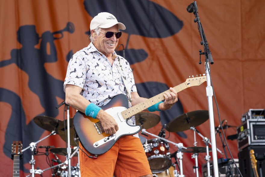 A older man with a ball cap and sunglasses, orange shorts and turquoise sweat bands on his wrist plays an electric guitar on stage while smiling down at the crowd.