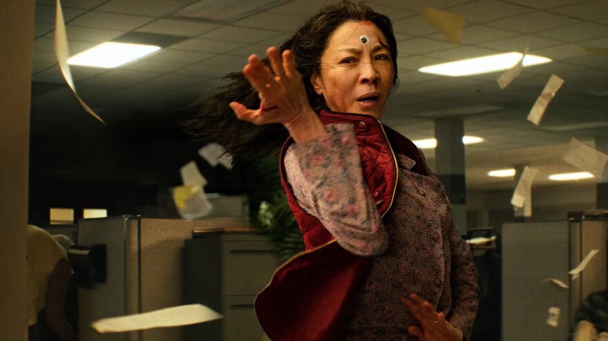 A woman with brownish skin, her hands in a dramatic martial arts pose, a googly eye on her forehead, as papers fly around her while she poses in an office environment.