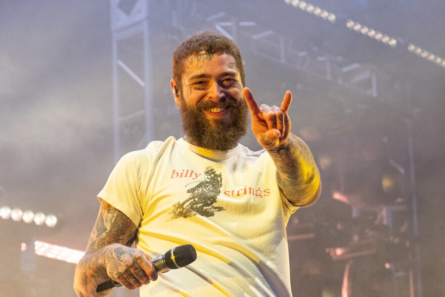 Music artist Post Malone smiles on stage while giving a rock hand sign.