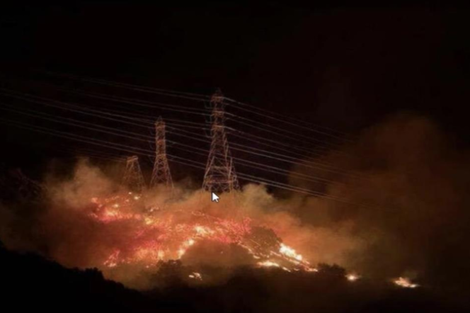 A burning hillside with powerlines within the flames is seen with smoke rising into the night sky.