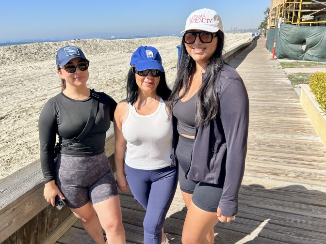Three female presenting persons stand on a wood walkway. All three wear sunglasses and baseball caps.