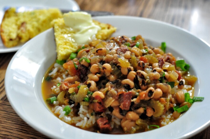 An up-close photograph of a shallow white bowl featuring a bed of white rice with saucy black-eyed peas ladled on top. The dish is accented with scallions and a slice of corn bread slathered with butter.