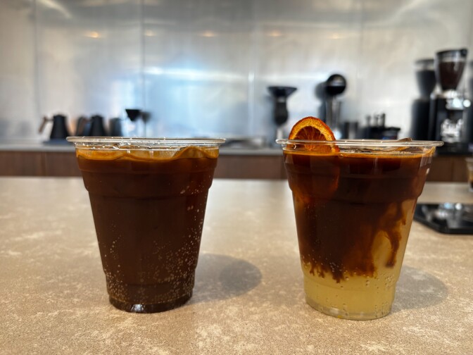 Two iced espresso drinks on the counter—one dark and fizzy, the other layered over citrus with a dried orange slice peeking from the rim in plastic cups.