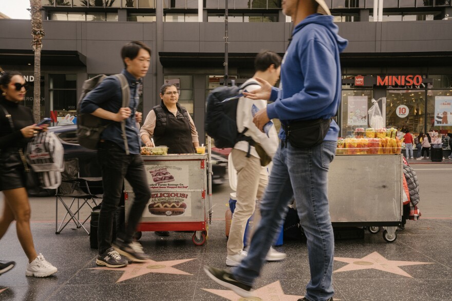 A busy street scene: Various pedestrians walk on the pink stars and black pavement of the Hollywood Walk of Fame, passing a woman who is standing at a hot dog cart.